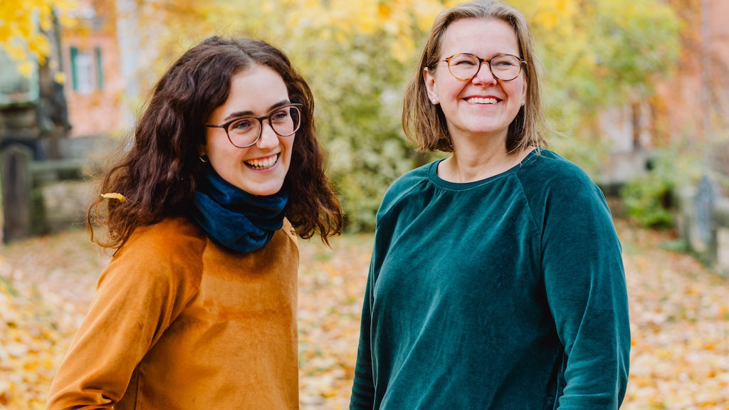Damen Nicki Pullover in Karamell und Dunkelgrün aus 100% Biobaumwolle von internaht - zwei fröhlich lachende Damen stehen in einem herbstlichen Park und tragen jeweils einen Nicki Pullover und einen Loop-Schal.