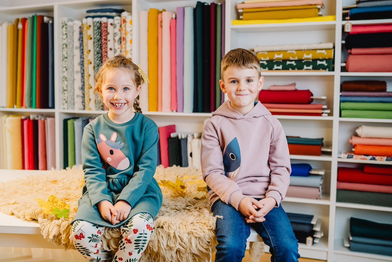 Kinder Pullover Jungen aus Bio Baumwolle. Ein Junge im beigefarbenen Hai-Hoodie und ein Mädchen im grünen Pferdekleid beides von internaht sitzen vor einem Stoffregal auf dem Tisch und lachen in die Kamera