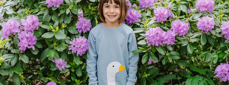 Kinder Pullover Mädchen aus Bio-Baumwolle von internaht, Mädchen steht vor lila blühenden Rhododendron-Busch und trägt einen himmelblauen Pulli mit Gans-Motiv und einen pinkfarbenen Stufenrock von internaht