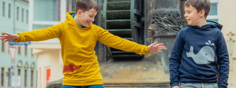 Kinderkleidung Jungen aus Bio-Baumwolle von internaht - zwei fröhliche Jungs stehen vor einem Brunnen, ein Junge trägt den senfgelben Dackelpullover mit Kapuze von internaht. Der andere Junge trägt den Wal Hoodie in Dunkelblau von internaht