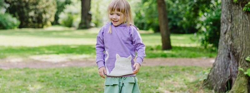 Kinderkleidung Mädchen aus Deutschland - ein Mädchen in einem grünen Park. Es trägt grünen Stufenrock und einen lilafarbenen Hoodie mit Katzenmotiv (alles von internaht)