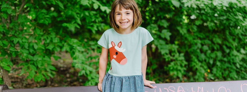 Schöne Mädchenkleider aus Biobaumwolle von internaht - ein Mädchen sitzt auf einer Parkbank und lacht in die Kamera. Es trägt in Kleid mit Pferdemotiv von internaht.