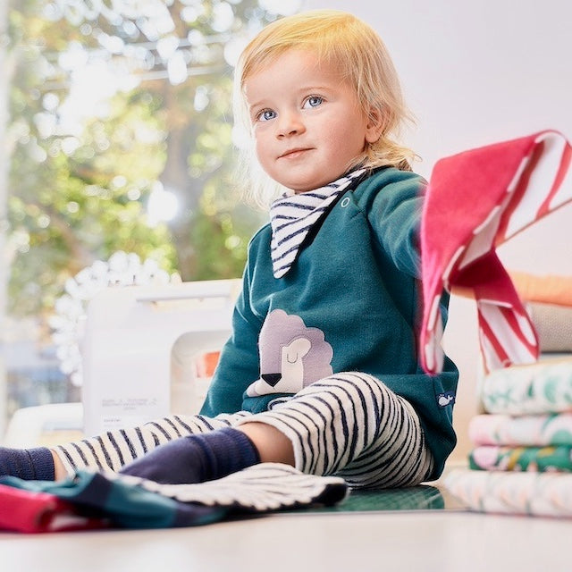 Fröhlich lächelndes Baby in einem grünen Baby Pullover aus 100% Bio Baumwolle mit Löwen-Motiv, passender gestreifter Babyhose und Baby Halstuch von internaht