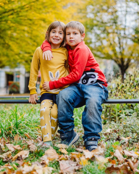 Fröhliche Kindermode für Jungen und Mädchen - Mädchen im senfgelben Sweater mit Schneehase und Junge mit Waschbär Sweater in Orange in einem herbstlichen Park