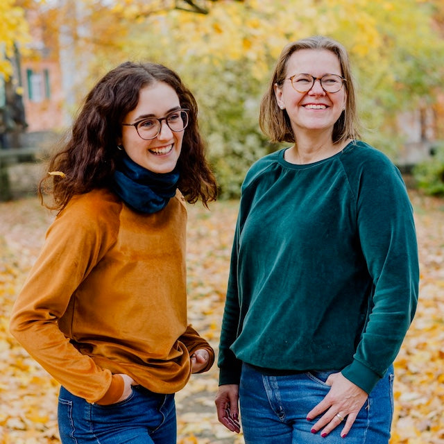 Damen Nicki Pullover in Karamell und Dunkelgrün aus 100% Biobaumwolle von internaht - zwei fröhlich lachende Damen stehen in einem herbstlichen Park und tragen jeweils einen Nicki Pullover und einen Loop-Schal.