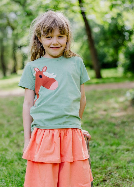 haltige Kindermode von internaht aus Dresden - Lächelndes Mädchen im Park trägt hellgrünes Kinder T-Shirt mit liebevollen Pferde-Motiv und orangefarbenen Stufenrock aus