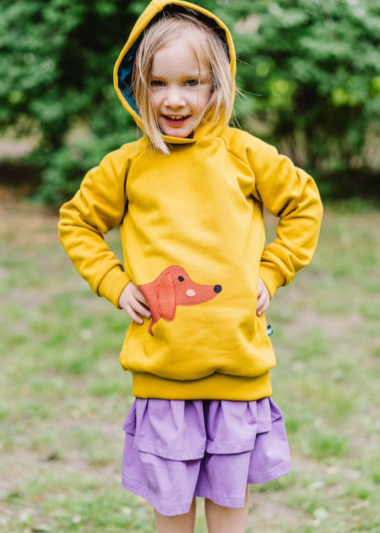 Fröhlich lachendes Mädchen im senfgelben Kapuzenpullover mit Dackel und lilafarbenen Stufenrock beides aus Bio Baumwolle von internaht - Nachhaltige Kindermode, die mitwächst und länger schön bleibt von internaht aus Dresden 